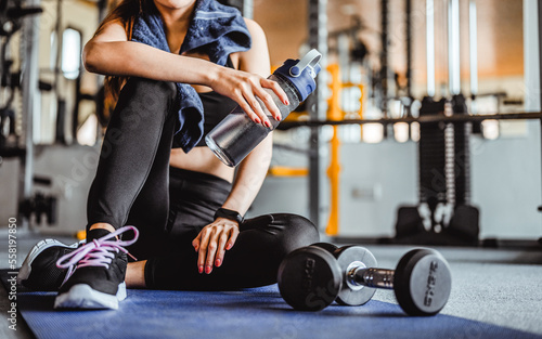 Fitness asian woman relaxing after exercise with drinking waters and dumbbell placed beside the gym.Relaxing after training.beautiful young woman looking away while sitting at Fitness gym.Fitness