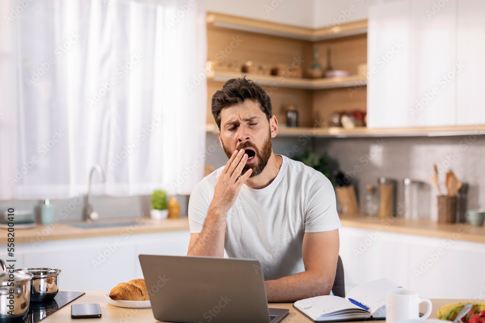 Sleepy tired bored handsome millennial caucasian guy with beard yawns, working at computer at table Sleepy tired bored handsome millennial caucasian guy with beard yawns, working at computer at table