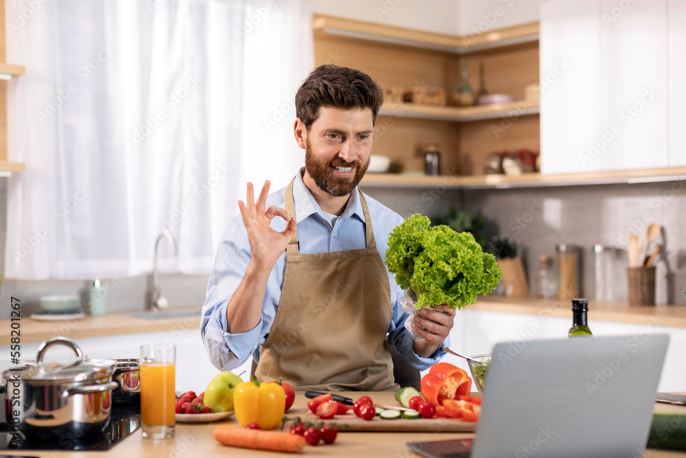 Obraz premium Glad millennial caucasian male cooking eat show salad and ok hand sign at table with vegetables and computer