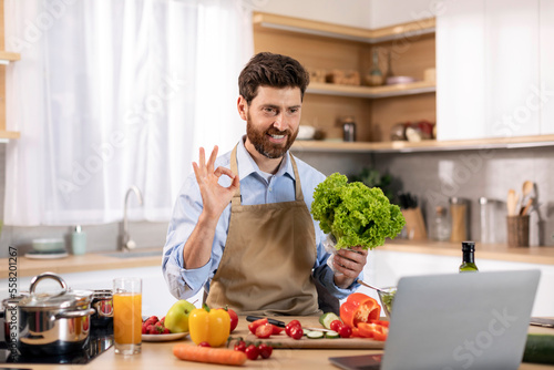 Glad millennial caucasian male cooking eat show salad and ok hand sign at table with vegetables and computer