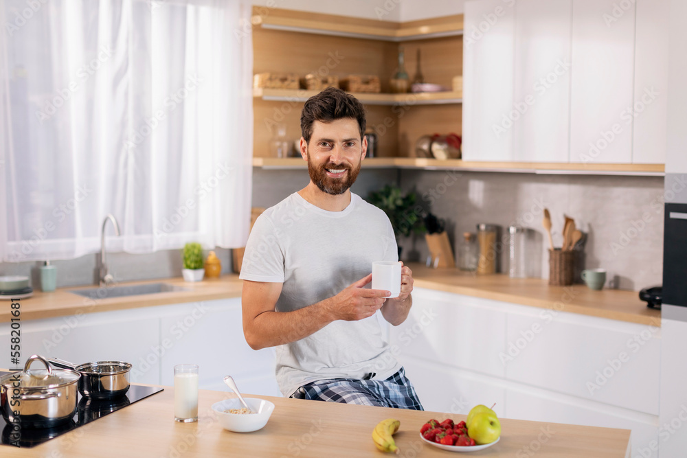 Smiling handsome millennial caucasian guy with beard in domestic clothes with cup of coffee enjoys good morning Smiling handsome millennial caucasian guy with beard in domestic clothes with cup of coffee enjoys good morning