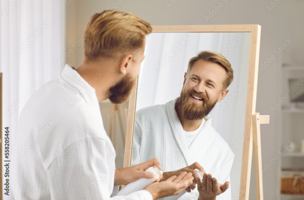 Fototapeta premium Grooming products for men. Happy bearded man with beard lotion in his hands enjoys morning routine of self care. Man in bathrobe looks at his reflection in the mirror. Morning male hygiene concept.