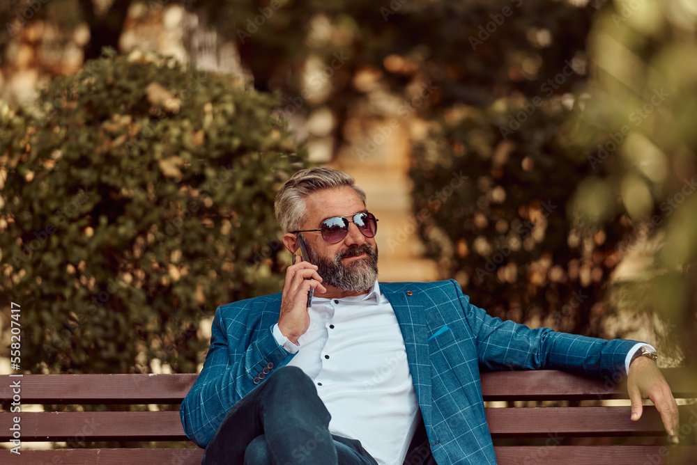 Modern senior businessman using smartphone for online meeting while sitting on bench Modern senior businessman using smartphone for online meeting while sitting on bench