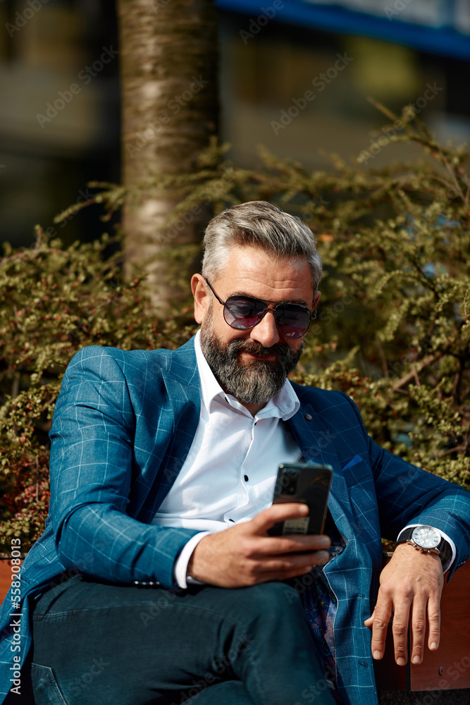 Modern senior businessman using smartphone for online meeting while sitting on bench Modern senior businessman using smartphone for online meeting while sitting on bench