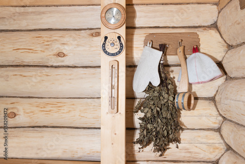 Bath accessories on a log wall, a birch broom, a sauna cap, a wooden bucket, sand clock and instruments for measuring humidity and temperature. Traditional old Russian spa concept