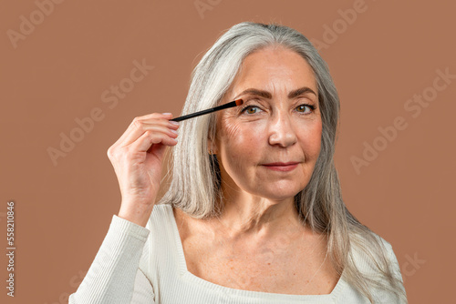 Serious pretty old caucasian female with gray hair applies cosmetics with brush isolated on brown background