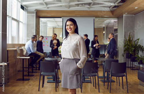 Fototapeta Naklejka Na Ścianę i Meble -  Portrait of friendly and successful business woman standing against background of colleagues in office. Millennial female leader or coach stands against background of people talking to each other.