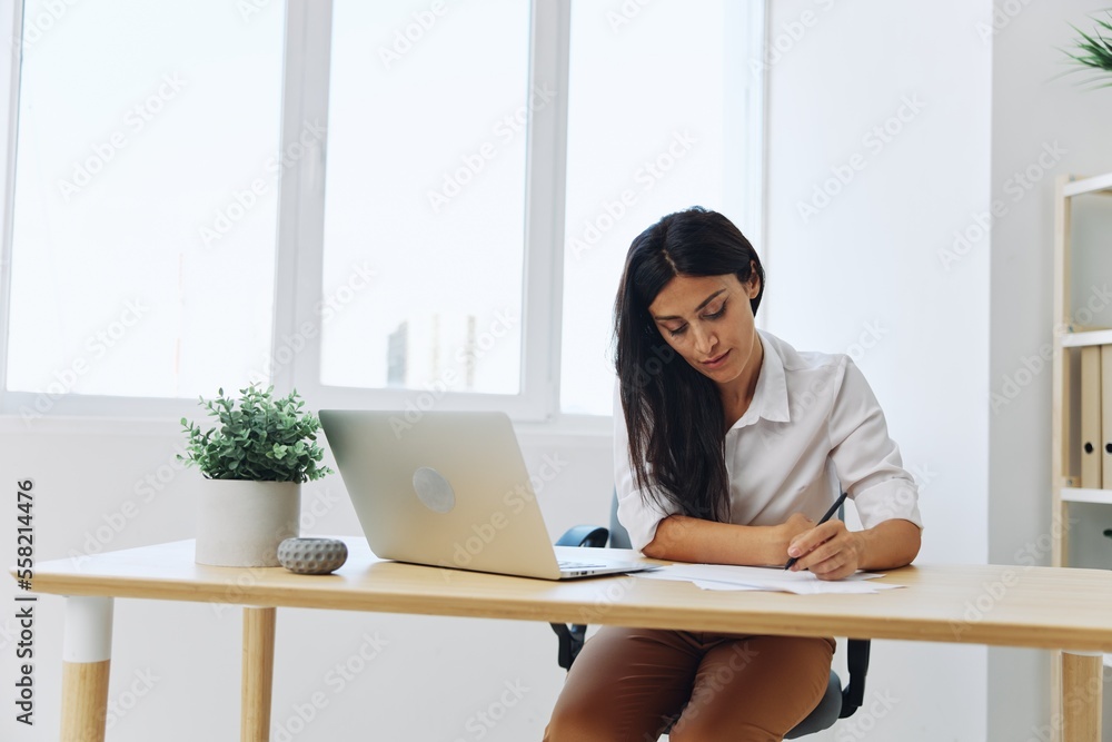 A woman works and learns by writing down text on paper with a pen and checking for errors, studying and teaching in college, business work at a desk with a laptop A woman works and learns by writing down text on paper with a pen and checking for errors, studying and teaching in college, business work at a desk with a laptop