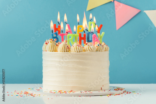 Birthday cake with white cream cheese frosting decorated with multicolored lit happy birthday text shaped candles on a blue background. Tradition of making a wish while blowing out candles on a cake