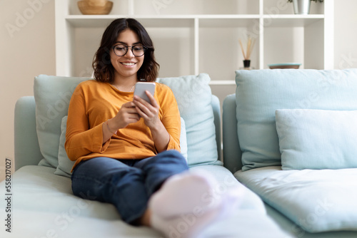 Beautiful Arab Female Wearing Eyeglasses Relaxing On Couch With Smartphone © Prostock-studio
