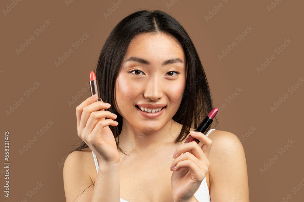 Beautiful asian woman holding two lipsticks in her hands and smiling, posing over brown studio background Beautiful asian woman holding two lipsticks in her hands and smiling, posing over brown studio background