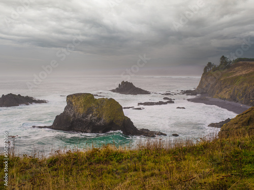 Large boulders in the ocean. A high rocky shore overgrown with yellow withered grass. There are gray storm clouds in the sky. Gloomy seascape. Nature, ecology, weather.