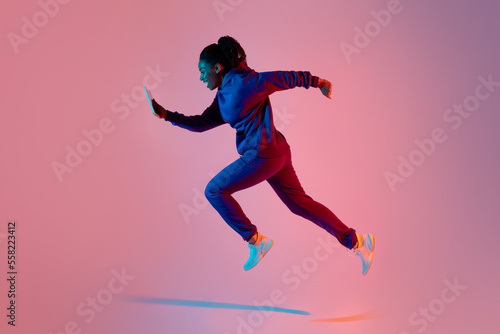 Full body length shot of modern sporty lady jumping or running using cellphone over pink neon background, side view