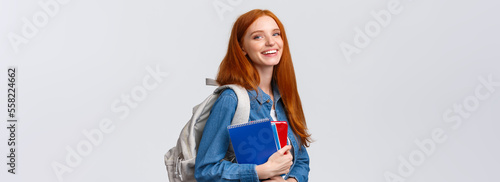 Teenagers, students and education concept. Cheerful lovely redhead female studying, going to univeristy or college, smiling carefree holding backpack and notebooks, standing white background