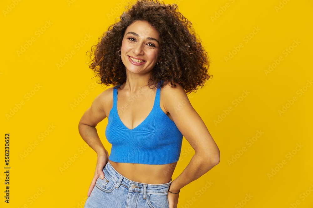 Woman with curly afro hair in a blue t-shirt on. yellow background signs with her hands, look into the camera, smile with teeth and happiness, copy space Woman with curly afro hair in a blue t-shirt on. yellow background signs with her hands, look into the camera, smile with teeth and happiness, copy space