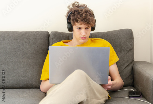 Cute smiling young teenager with laptop and headphones on the couch at home. Boy makes homework with a computer. Distance, online education for children