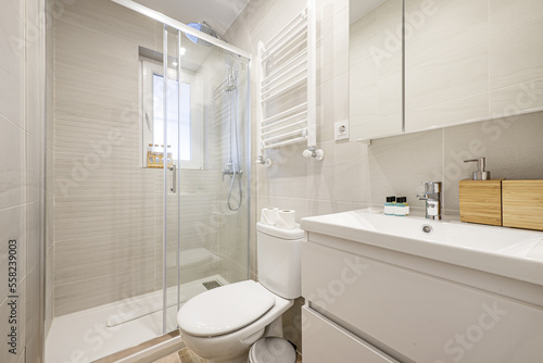 Small bathroom with white porcelain sink on wooden chest of drawers with glass-door cabinets and glass-enclosed shower stall
