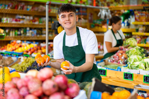 Obraz na płótnie Portrait of cheerful young male in apron selling organic tangerines and fruits i