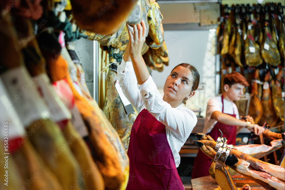 Skilled interested young girl, owner of butcher shop selling delectable dried Iberian jamon working at counter