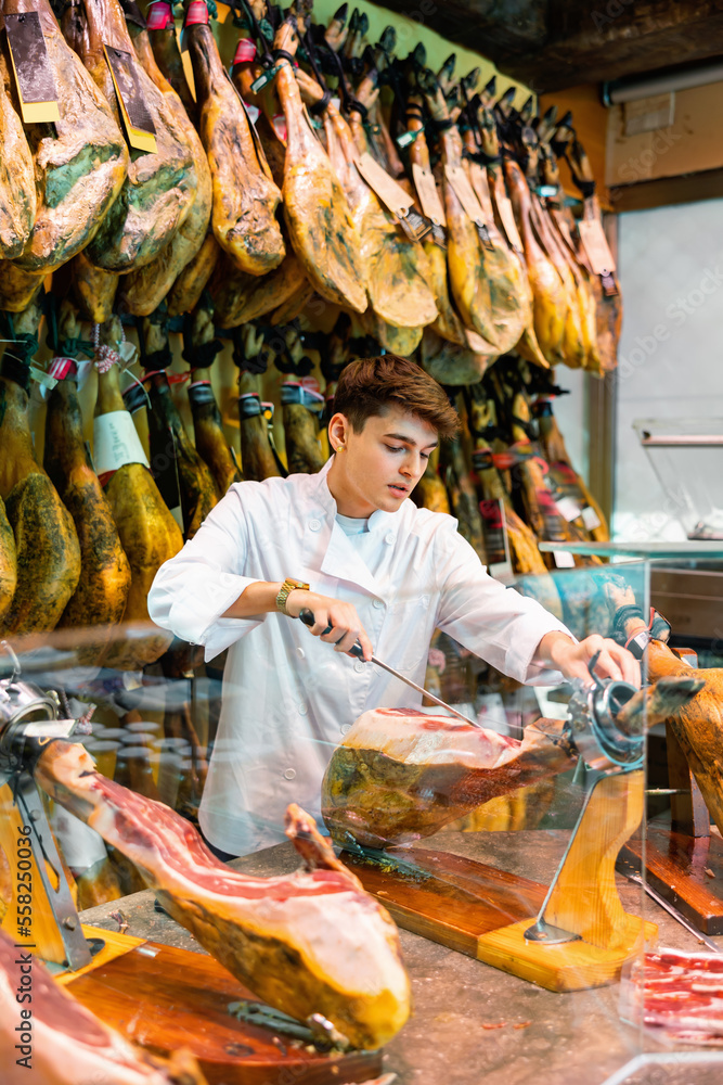 Young man working in jamoneria, standing at counter and cutting ham with special knife.