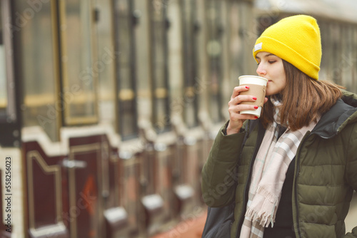Travel and coffee concept: portrait of beautiful young woman drinking hot beverage at street of European city. Model wearing green coat, white and beige scarf. Text space. Outdoor shot