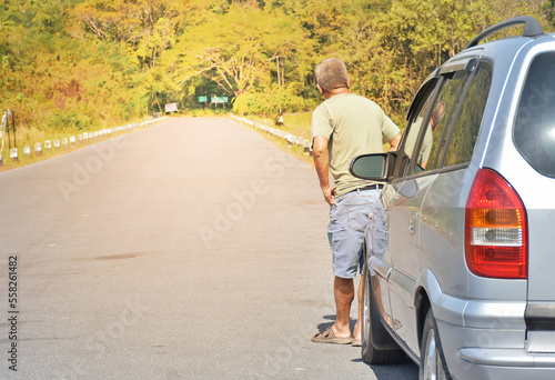 Back view of Middle-aged Asian man stood standing alone in front of car and looked out to end of road and waiting for help in case of passing car between travel. Vehicle check up for travel concept.