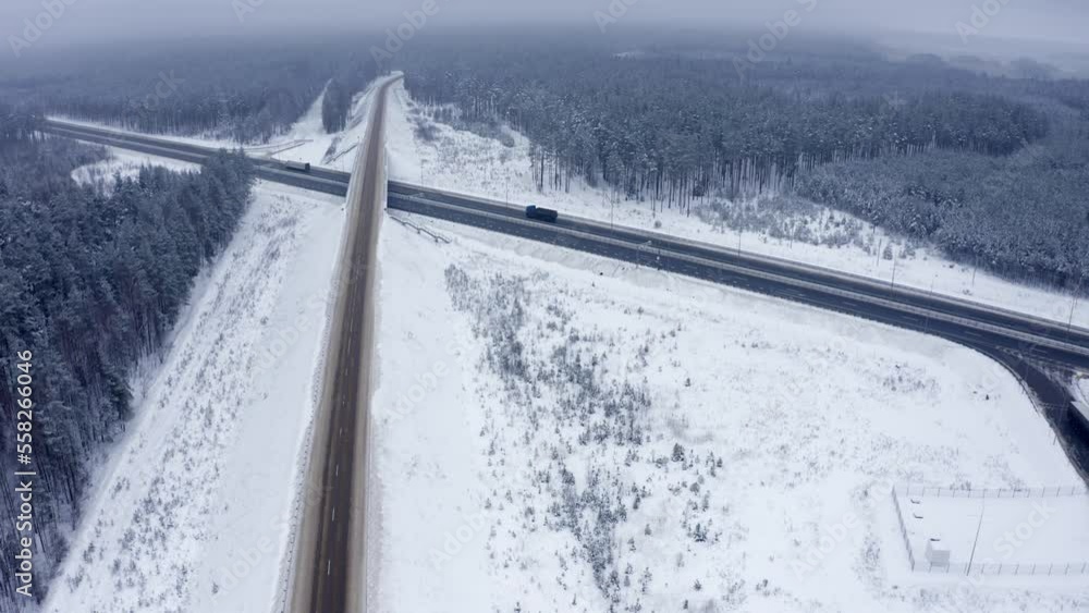 Several trucks drive along the freeway under a bridge past a parking lot with parked cars and in the middle of a snowy forest. Side view. In winter, two trucks drive along the road. Several trucks drive along the freeway under a bridge past a parking lot with parked cars and in the middle of a snowy forest. Side view. In winter, two trucks drive along the road.