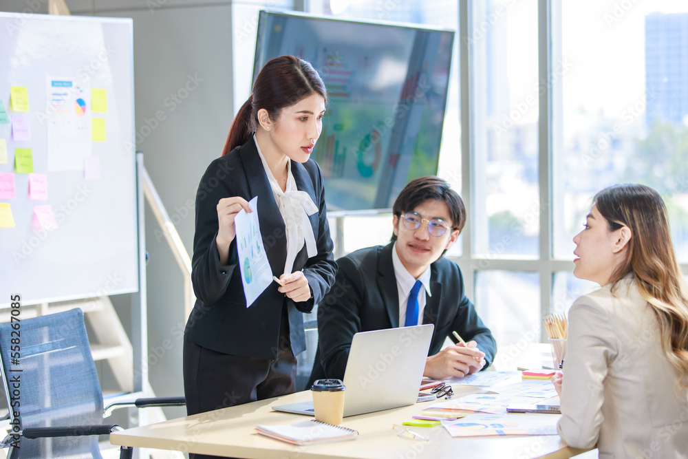 Fototapeta premium Asian professional successful female businesswoman lecturer presenter in formal suit standing smiling at graph chart data answer questions to male colleague in company office meeting room.