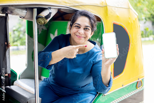 Happy mature indian woman enjoying rickshaw ride showing blank smart phone display screen for advertisement.