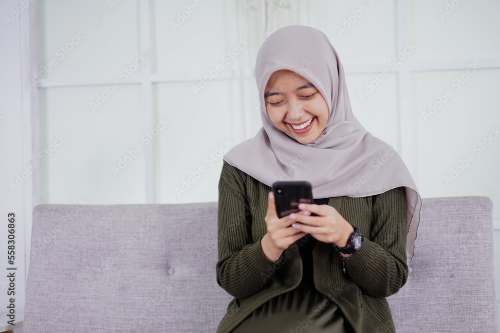 Fototapeta premium A beautiful Asian woman in hijab smiles cheerfully sitting on a sofa using a mobile phone, receiving good news