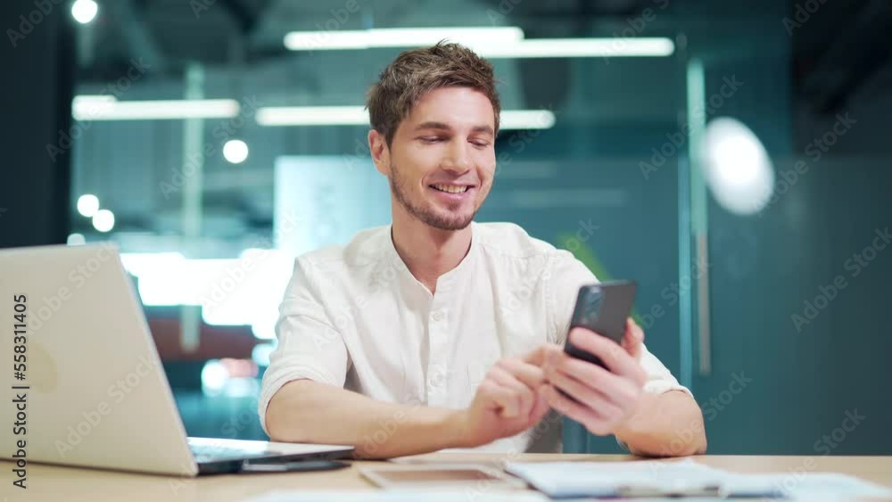 Happy employee using smartphone at work in modern office. Satisfied caucasian manager using a mobile phone while working, browsing the Internet, answering email, texting with friend or family, smiling Happy employee using smartphone at work in modern office. Satisfied caucasian manager using a mobile phone while working, browsing the Internet, answering email, texting with friend or family, smiling
