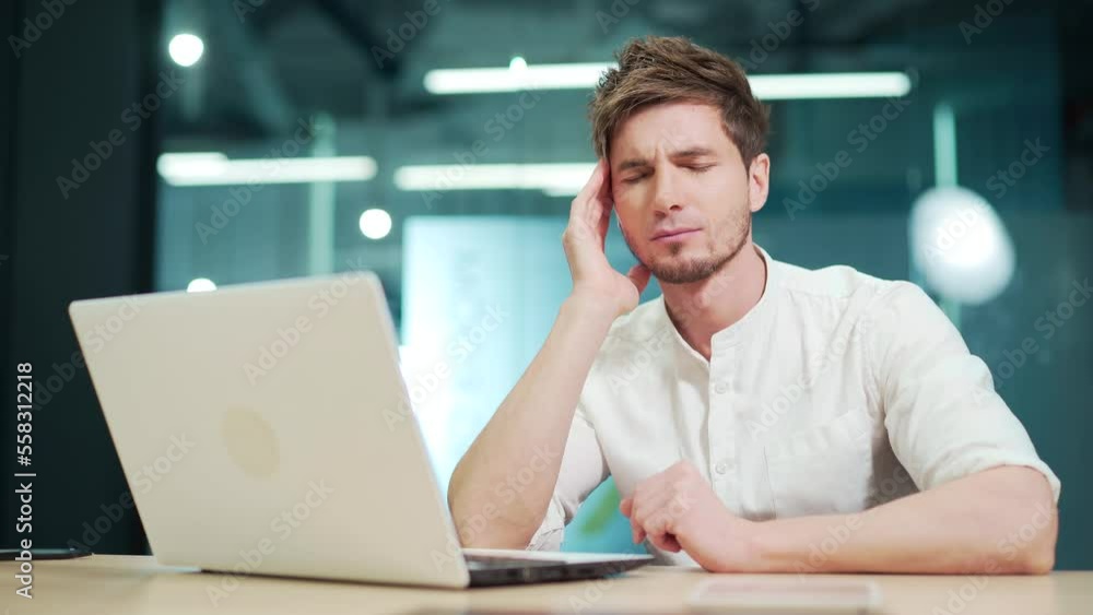 Handsome young man feels tired and has a headache at work in the office. Caucasian male massages temples with his hands. Overworked stressed businessman feeling pain sitting at desk with laptop indoor Handsome young man feels tired and has a headache at work in the office. Caucasian male massages temples with his hands. Overworked stressed businessman feeling pain sitting at desk with laptop indoor