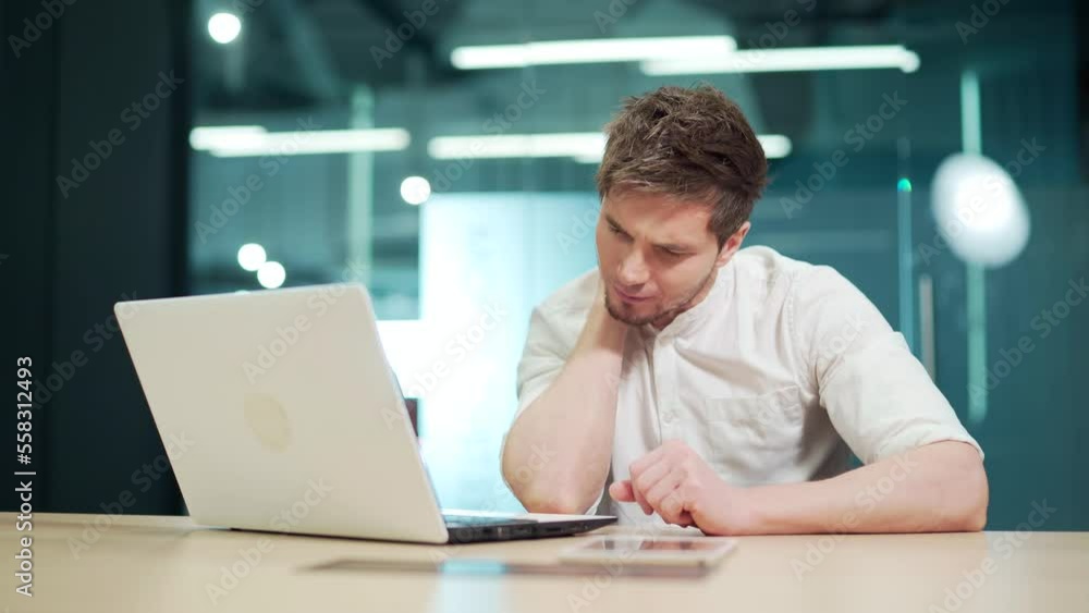 A young man feels pain in his neck while working in the office on a laptop. A male massages his neck, Feeling discomfort in the muscles from a long sedentary lifestyle. Rubs his shoulder with his hand A young man feels pain in his neck while working in the office on a laptop. A male massages his neck, Feeling discomfort in the muscles from a long sedentary lifestyle. Rubs his shoulder with his hand