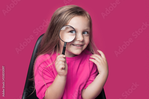Little girl sitting on chair, holding magnifying glass and looking at camera with curious expression, wearing pink background, isolated over pink background.