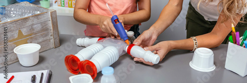 Unrecognizable teacher helping to girl make recycled toy robot with hot melt glue gun in ecology classroom