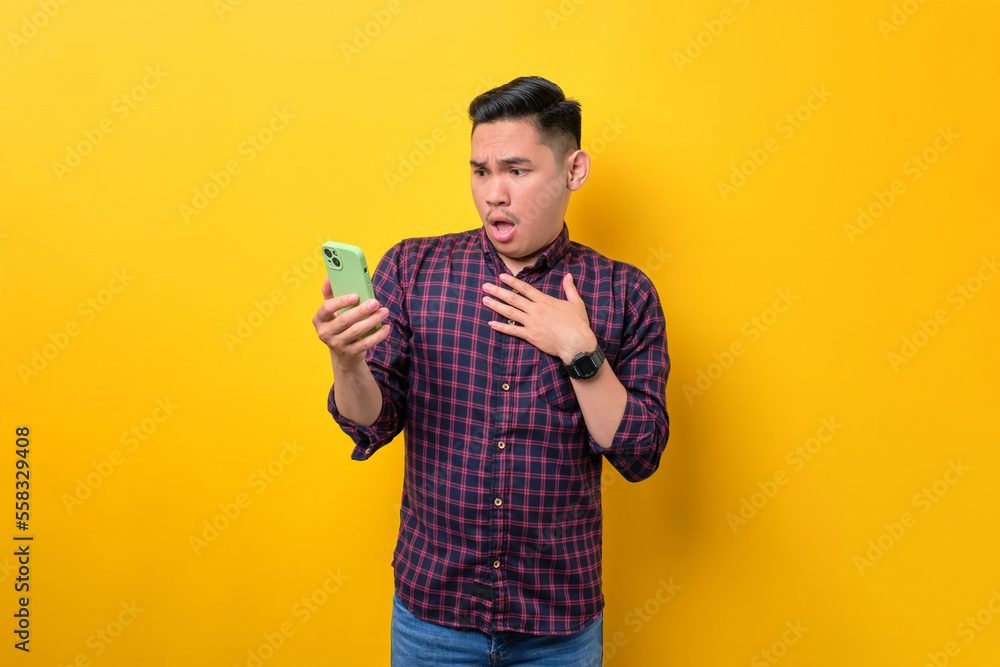 Surprised young Asian man using smartphone, reading special offer isolated over yellow background Surprised young Asian man using smartphone, reading special offer isolated over yellow background