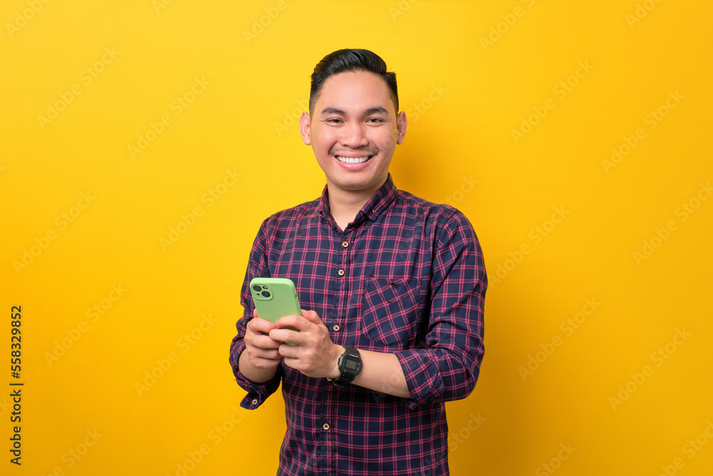 Cheerful young Asian man holding smartphone and smiling at camera isolated over yellow background Cheerful young Asian man holding smartphone and smiling at camera isolated over yellow background