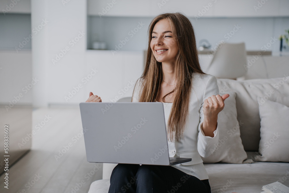 Successful young female entrepreneur talks via internet  clenched fists like a winner, smiling making video call at home, sits on couch, using laptop. Business and remote education. Great deal.