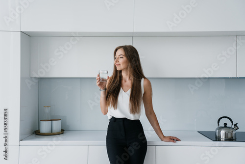 Satisfied fit businesswoman in elegant clothes standing at kitchen holds glass of water looks aside smiles. Attractive Italian female satisfied by healthy lifestyle. Successful entrepreneur dreaming.