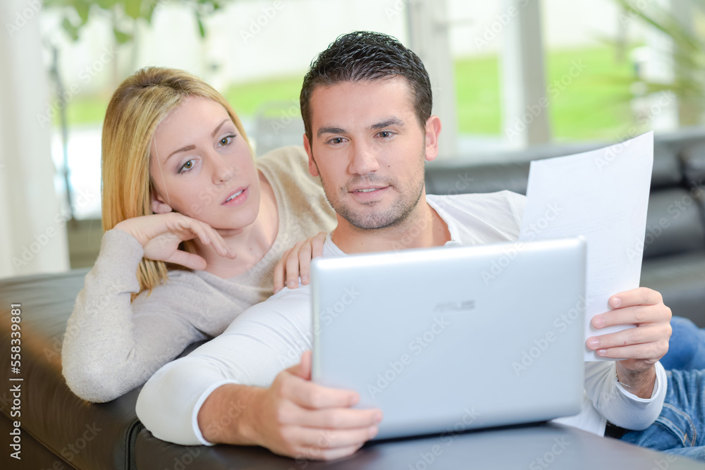 Couple looking at computer, holding sheet of paper Couple looking at computer, holding sheet of paper