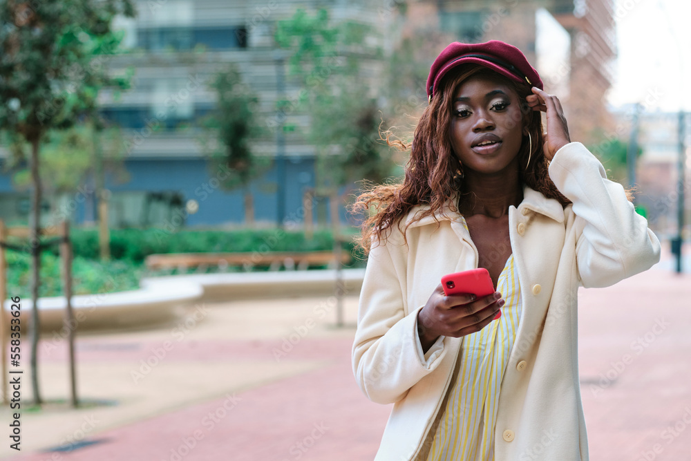 Stylish woman looking at camera while holding a mobile phone outdoors on the street.
