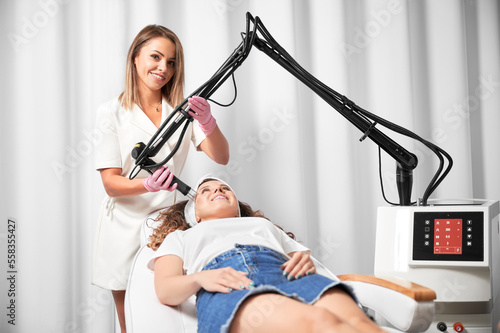 Smiling woman lying on daybed and receiving facial skincare treatment in beauty salon. Female doctor cosmetologist using erbium ablative laser machine while performing resurfacing procedure.