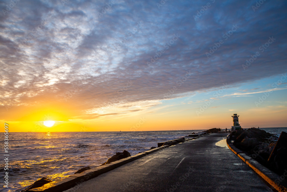 path to lighthouse on zuidpier IJmuiden at sunset with cloudy sky and sun above horizon