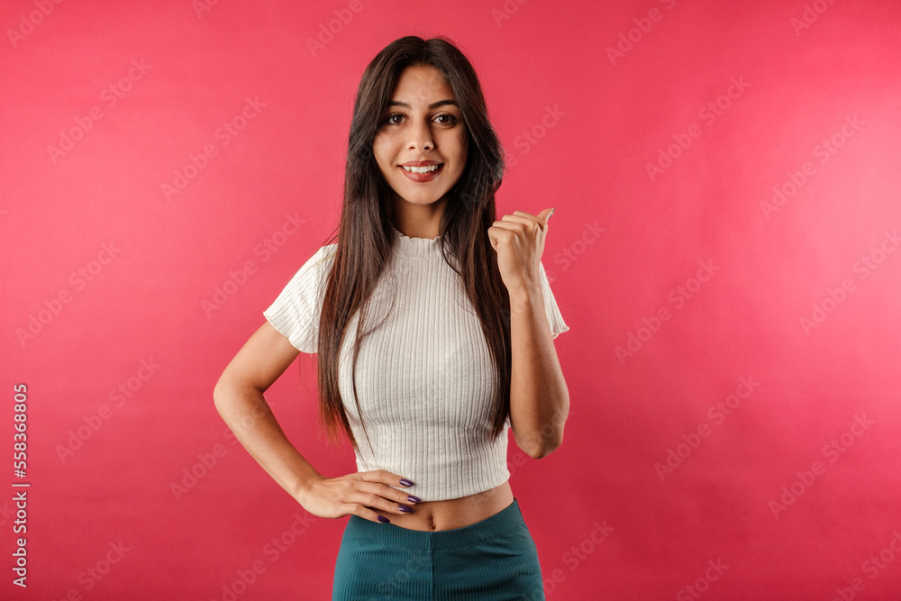 Happy young woman wearing white ribbed crop isolated over red background smiling and pointing to the copy space with her thumb while looking at the camera Happy young woman wearing white ribbed crop isolated over red background smiling and pointing to the copy space with her thumb while looking at the camera