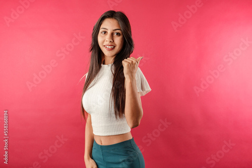 Cute caucasian woman wearing ribbed crop isolated over red background pointing thumb finger back behind on workspace area copy space. Looking at the camera.