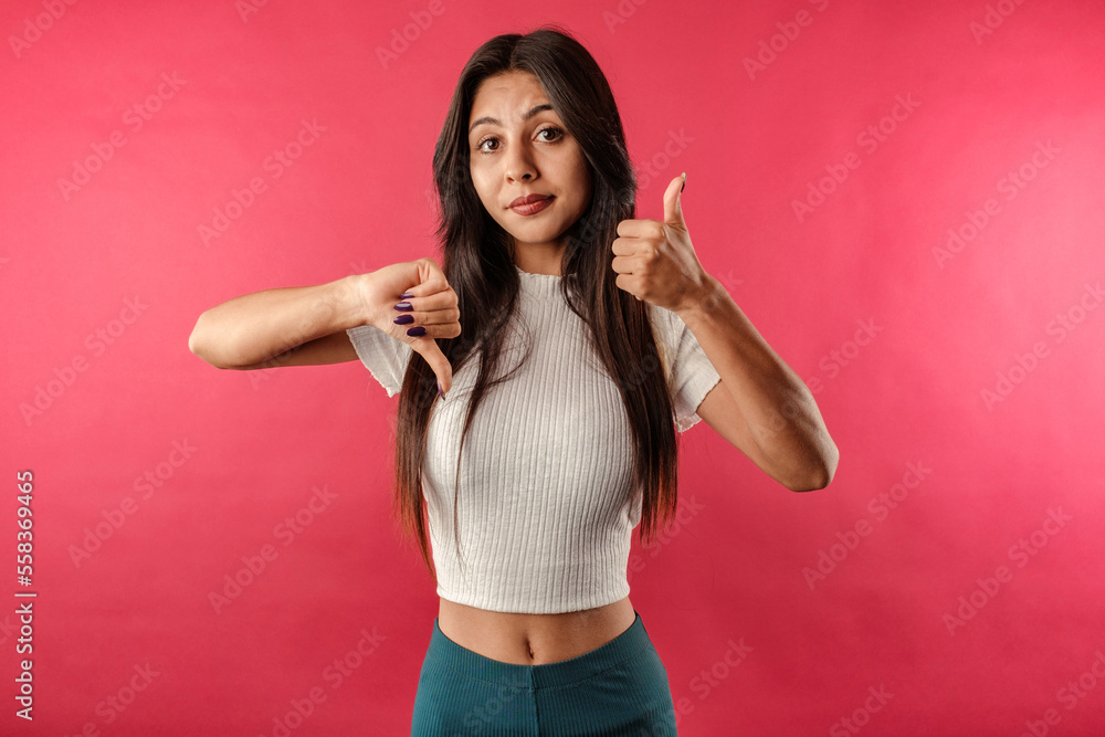 Young beautiful woman wearing ribbed crop isolated over red background doing thumbs up and down, disagreement and agreement expression. She looks unsure.