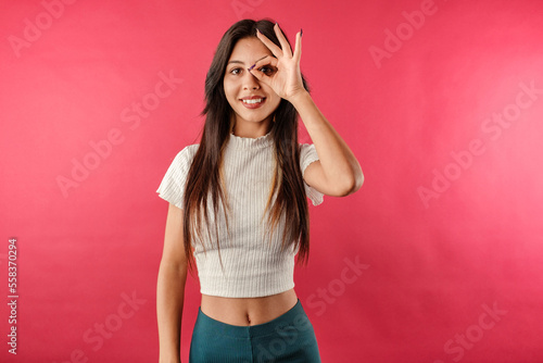 Happy brunette woman wearing casual top isolated over red background looking through okey sign. Happy, confident and everything is fine. Face expressions, emotions, and body language.