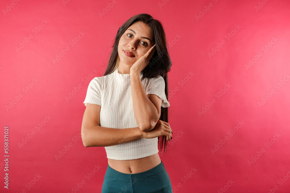 Young dark-haired woman wearing ribbed crop isolated over red background looking reluctant and unamused at camera, leaning face on hand. Listening boring person. Listens to lies and does not believe. Young dark-haired woman wearing ribbed crop isolated over red background looking reluctant and unamused at camera, leaning face on hand. Listening boring person. Listens to lies and does not believe.