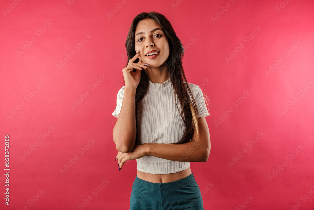 Cute brunette woman wearing white ribbed crop isolated over red background enjoys talking and laughs a lot. He touches his chin with his hand and likes it. Cute brunette woman wearing white ribbed crop isolated over red background enjoys talking and laughs a lot. He touches his chin with his hand and likes it.