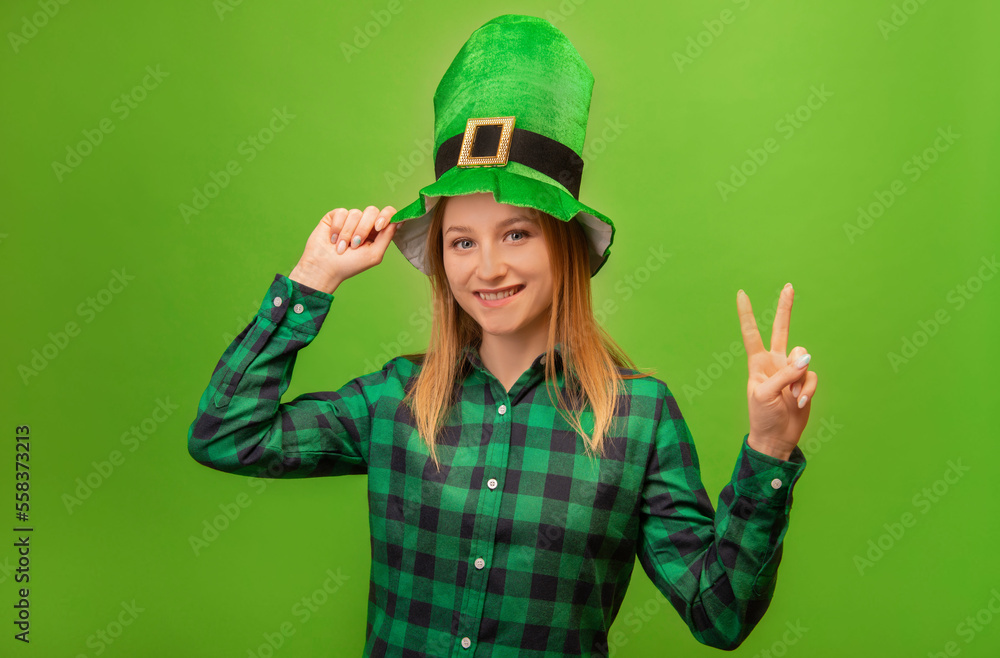 St Patricks Day leprechaun girl in green festive irish hat and checkered plaid shirt showing peace gesture v sign with two fingers and looking at camera isolated on green background. St Patricks Day leprechaun girl in green festive irish hat and checkered plaid shirt showing peace gesture v sign with two fingers and looking at camera isolated on green background.